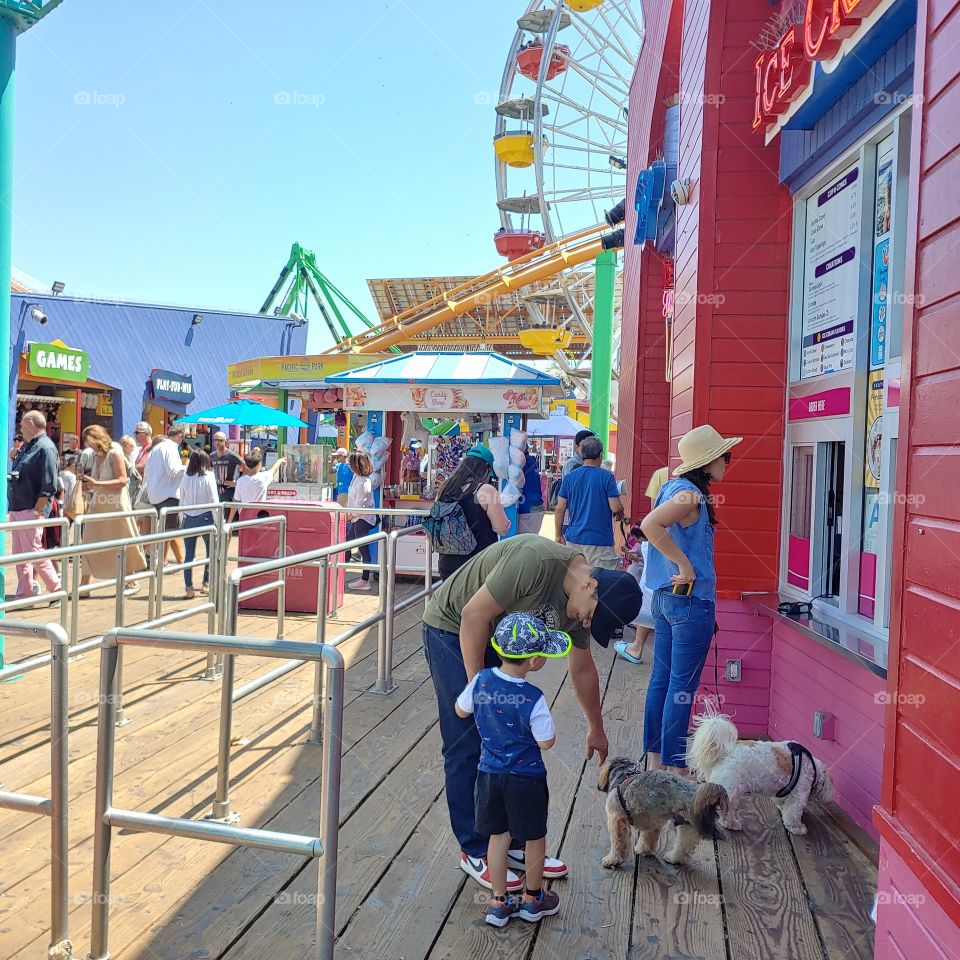 The photo shows a weekend carnival experience at Santa Monica Pier CA with a kid and his grandfather petting a dog while buying ice cream