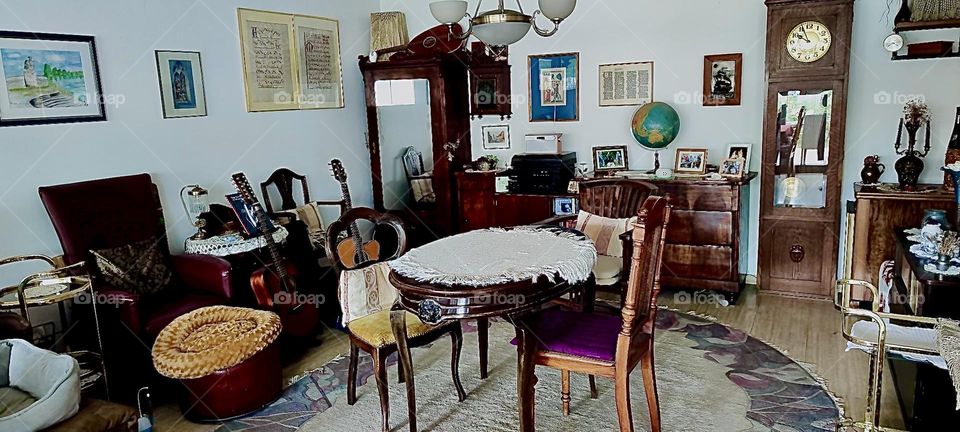 Antique furniture fill this part of the living room in the beautiful house in “Bavaria”, Germany. A table with chairs, a tall glass door cabinet, chest w drawers, globe, artwork, musical instruments make the decor complete. 2023. Hypnotic Productions