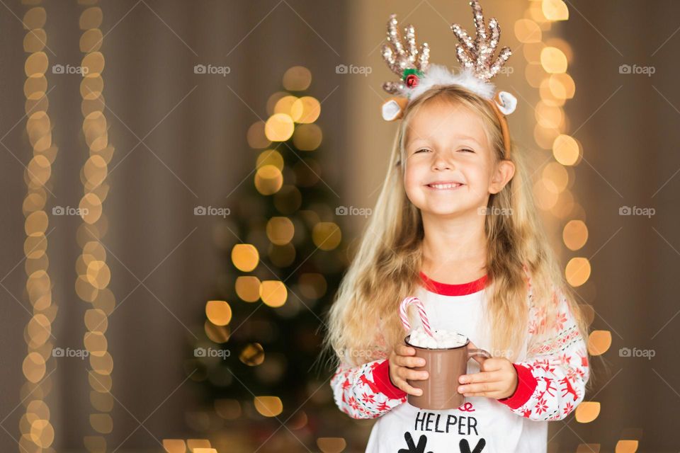 Little girl wearing Christmas pajamas and drinking hot cocoa with marshmallow at home 