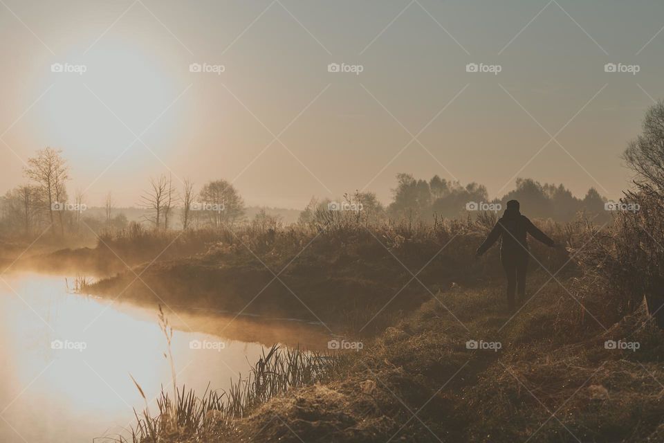 Woman walking through a meadow by a pond in the foggy morning. Sun rising above field and pond flooded with fog in the morning. Real people, authentic situations