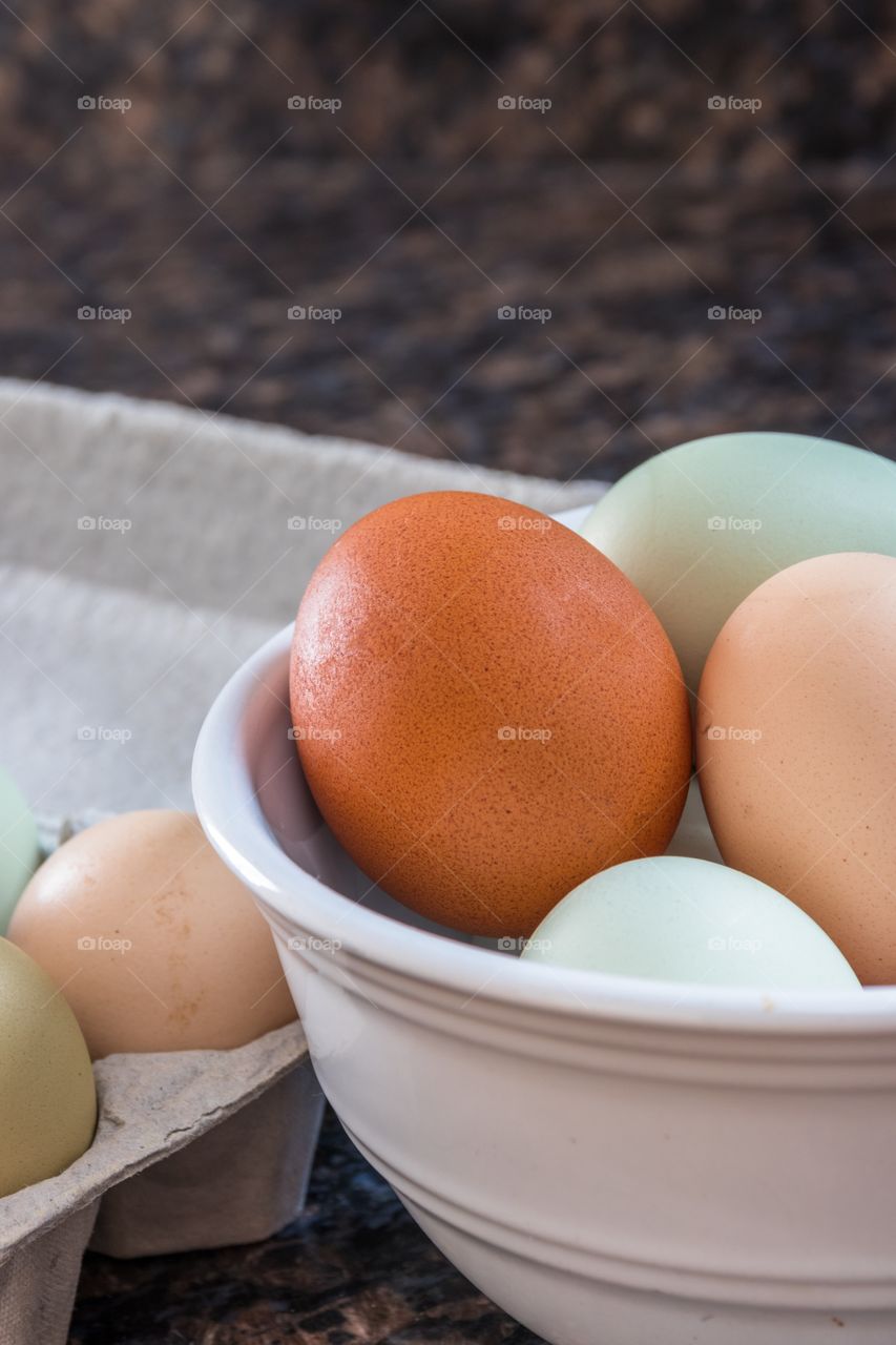 Vertical close up photo of multicolored eggs in a white bowl on a kitchen counter with more eggs in a grey carton to the side