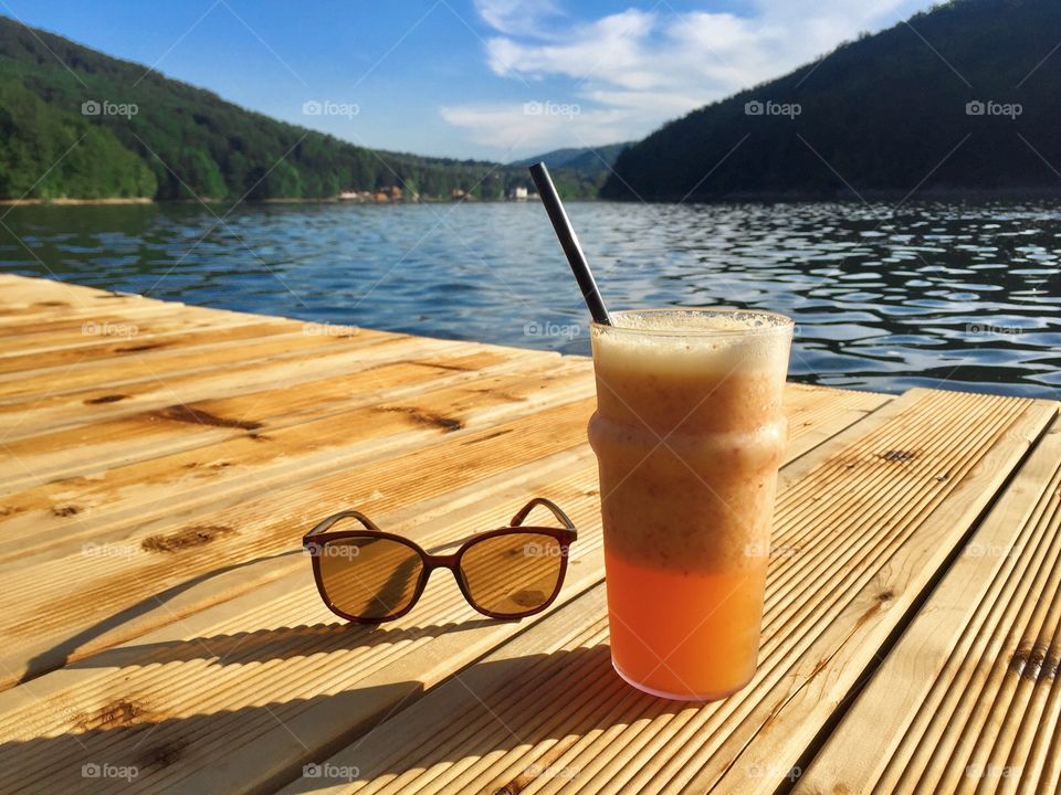 Glass of orange smoothie with drinking straw with a pair of sunglasses on a rustic wooden pontoon near the lake