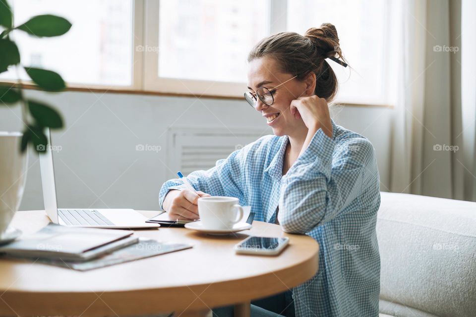 Young smiling woman in blue shirt using laptop and doing notes in room