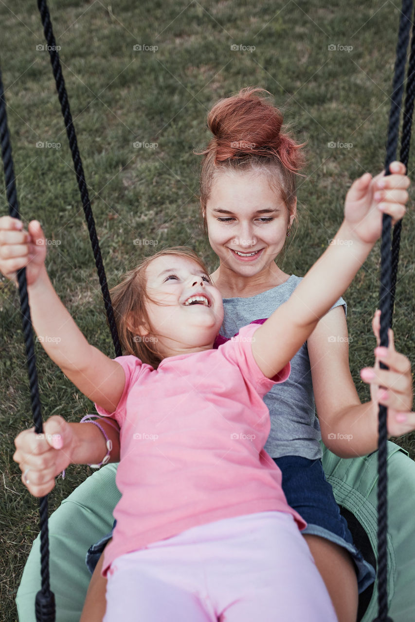 Teenage girl playing with her younger sister in a home playground in a backyard. Happy smiling sisters having fun on a swing together on summer day. Real people, authentic situations