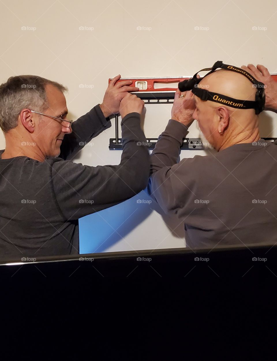 Men remodeling & figuring a plan to wall mount an LED TV. There's a level on top, keeping the mount in place. First they must find the wall studs. One wears a headlamp to keep his hands free to use tools.