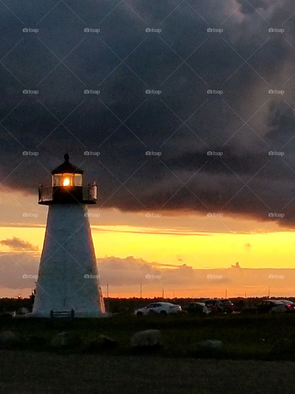 Sunsets & Sunrises of the USA! Lighthouse in New England lit, behind are dark storm clouds with a sunrise/sunset in background. Daylight is visible low in the sky on the horizon. Boulders surround the area of the lighthouse.