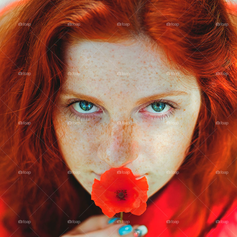 Close up portrait of a young redhead lady with freckles and blue eyes sitting outside with red poppies in her hands.