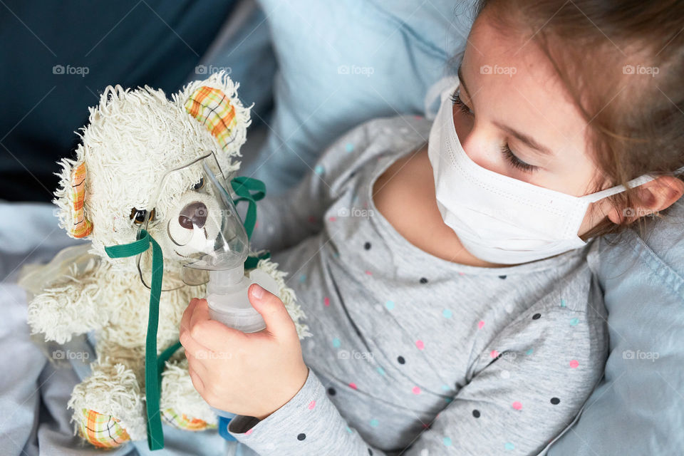 Sick child recovering in bed. Little girl playing by applying medical inhalation treatment with nebuliser to her teddy bear