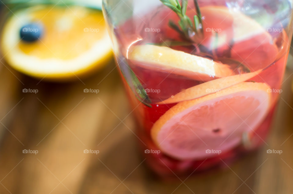 Closeup of healthy homemade flavored water made with citrus and fresh summer blueberries, raspberries, lemon, orange and rosemary in glass half full on wood background with ingredients 