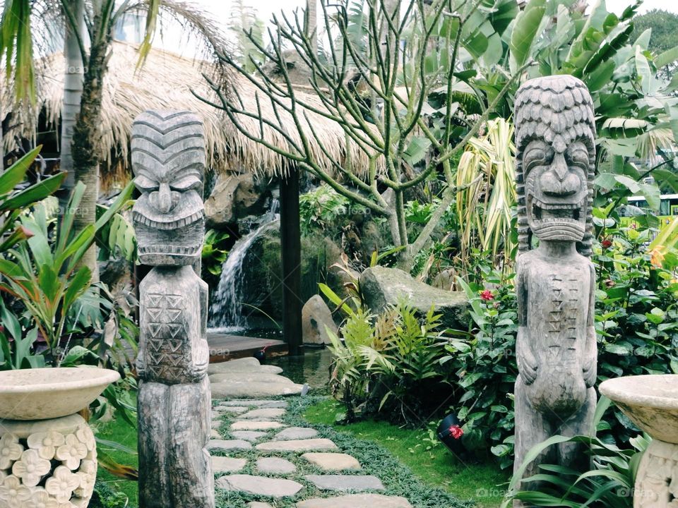 A dreamy garden with a winding path leading to a quaint hut adorned with intricate stone carvings and a thatched roof. A gentle waterfall flows in the background, surrounded by lush tropical plants, creating a serene and enchanting atmosphere.