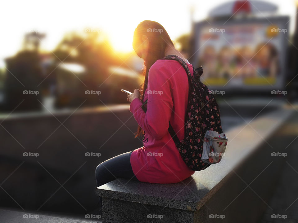 Young modern woman-student with a backpack using mobile phone at sunset time in the city 