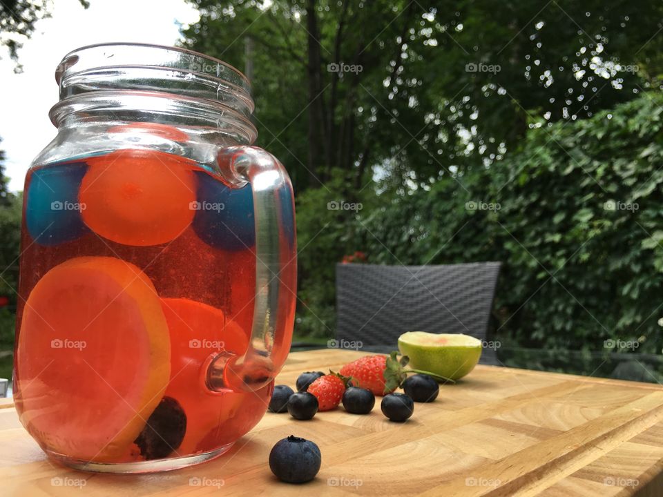 Glass of homemade flavored water with raspberries, lemon, strawberry and blueberry in garden on patio table refreshing healthy hydrating summer drink with plastic ice cube balls