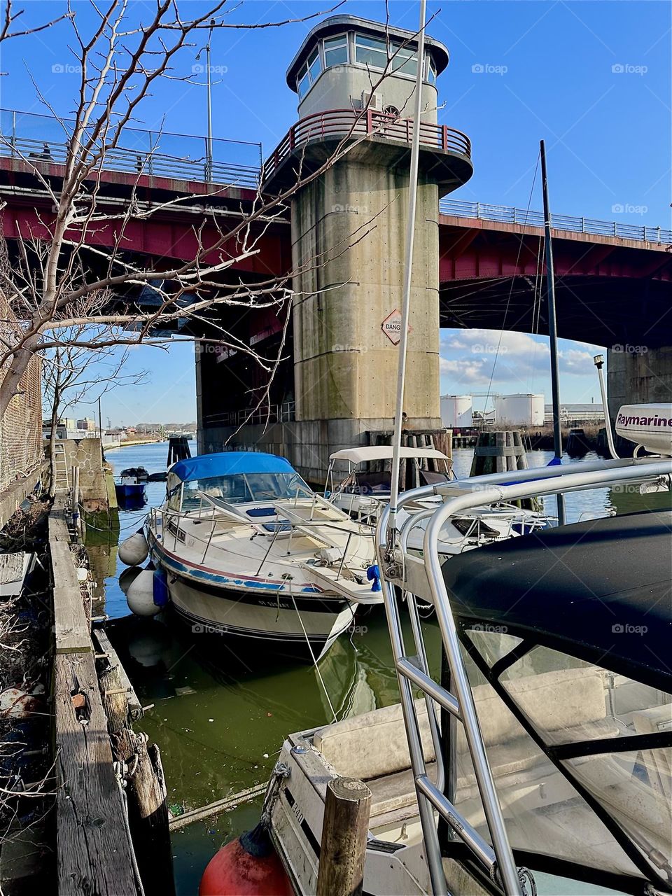 This is “Newtown Creek” by the “Pulaski Bridge” in LIC, Queens seen from the narrow strip of land that holds several wooden planks that various  sailboats and motorboats are tied to up to three deep. 2023. Hypnotic Productions