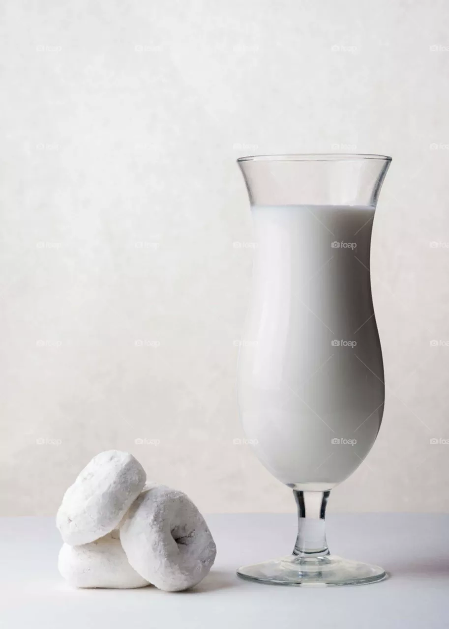 white milk in a glass next to mini white powder doughnuts on a white table in front of a white background