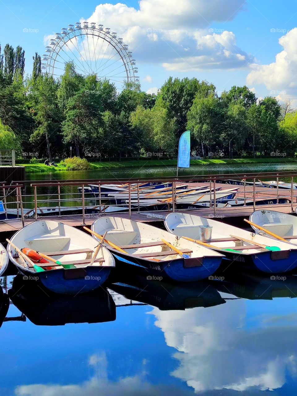 Summer. A park. A pier where pleasure boats are moored. The clear water reflects boats and the sky with white clouds. Behind the green trees is a Ferris wheel attraction.