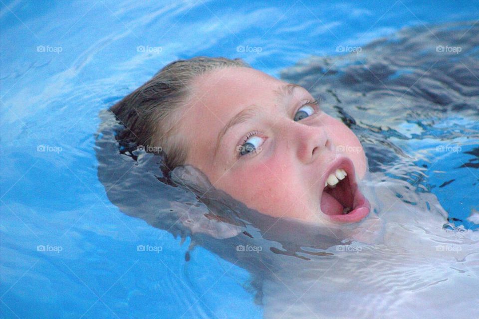 A goofy young girl getting a mouthful of pool water as she floats on her back. 