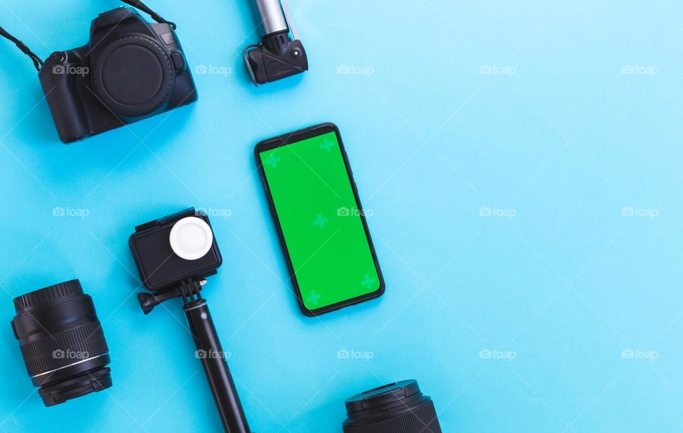 Photographer's set of cameras, lenses, selfie sticks and a smartphone with a green screen lie on the left on a blue background with copy space on the right, flat lay close-up.