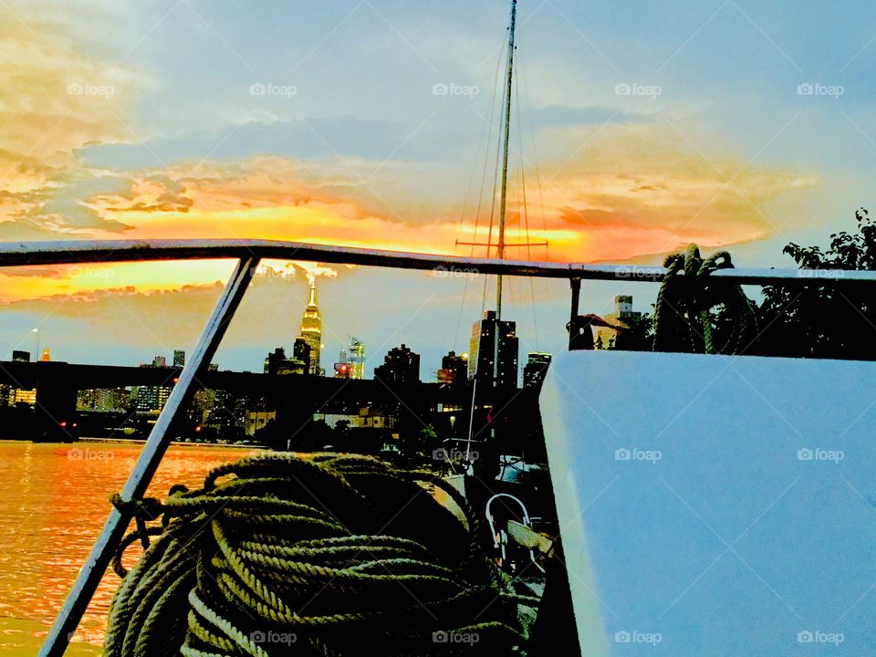 Aboard the “Salvation”, our sailboat on the front deck under a colorful evening sky with the ocean, Long Island City, Queens, NY, a silhouette of Manhattan and the Empire State Building in the background. Photo from 2018. Hypnotic Productions
