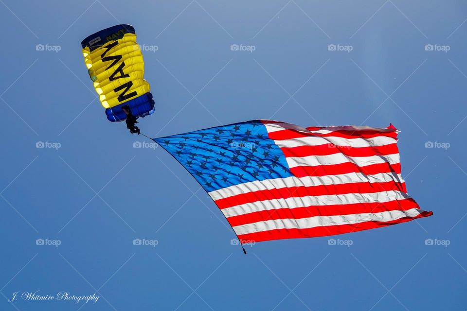 A member of the United States Navy skydiving team has a very large American flag tied to his body as he descends over a crowd