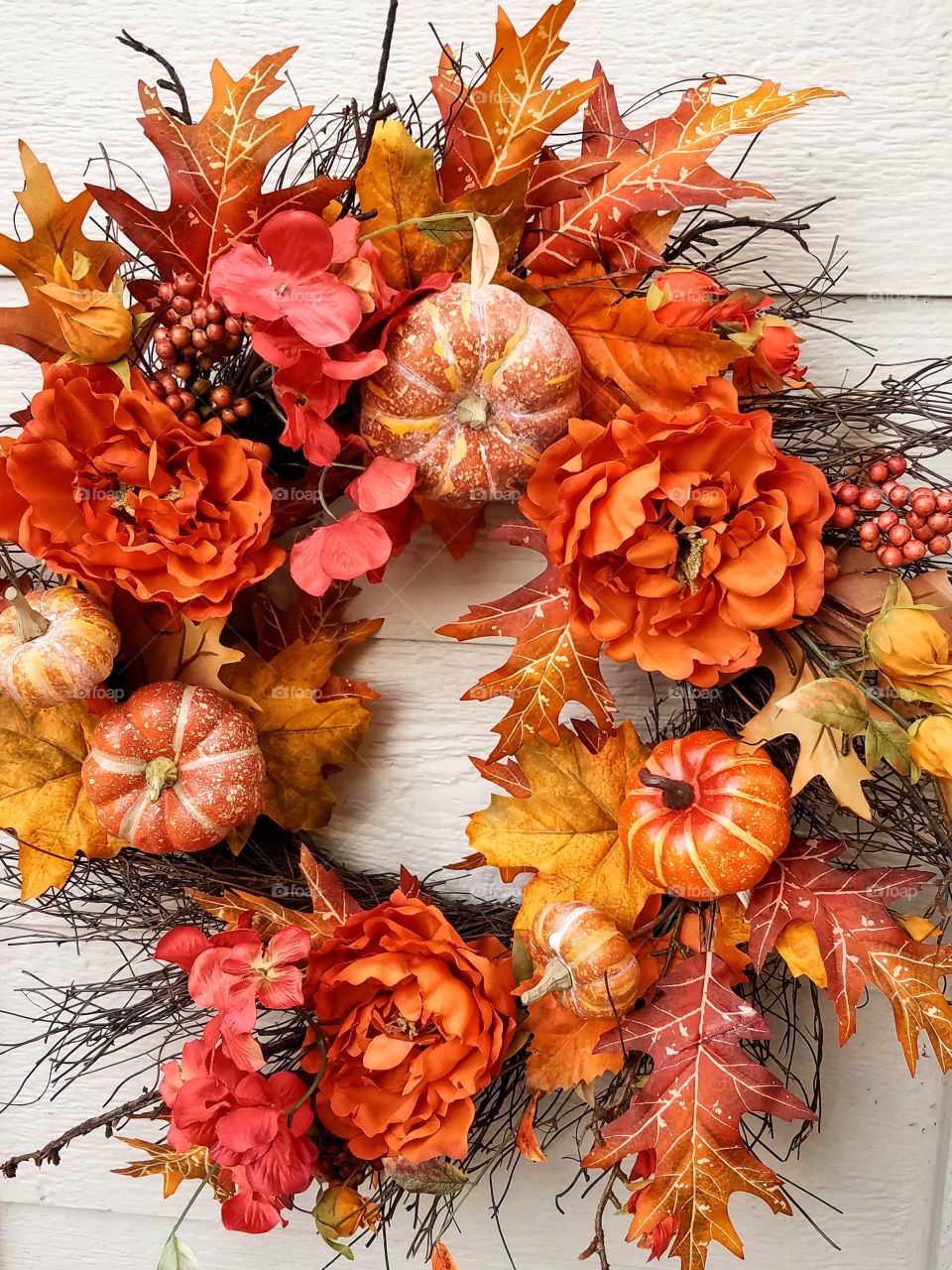 a colorful, orange and yellow, autumn,  door wreath made of small round gourds, flowers, and oak leaves against an off-white wall