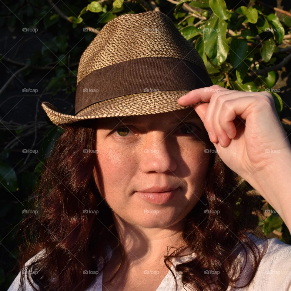 Women with natural and minimal makeup standing outdoors outside with wet hair. Women with red hair and green eyes standing next to tree taking a selfie. Sunlight shine on women hair and skin and making shadows. Women wearing a hat.