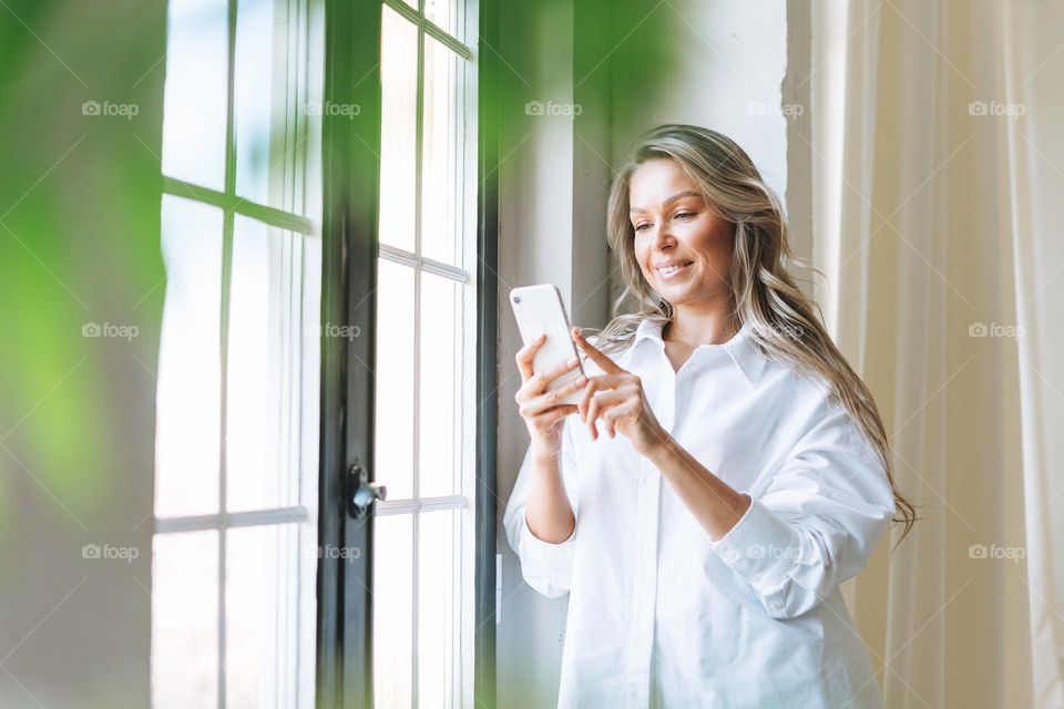 Young smiling blonde woman with long hair in white shirt using smartphone in the bright modern office, woman send message by smartphone