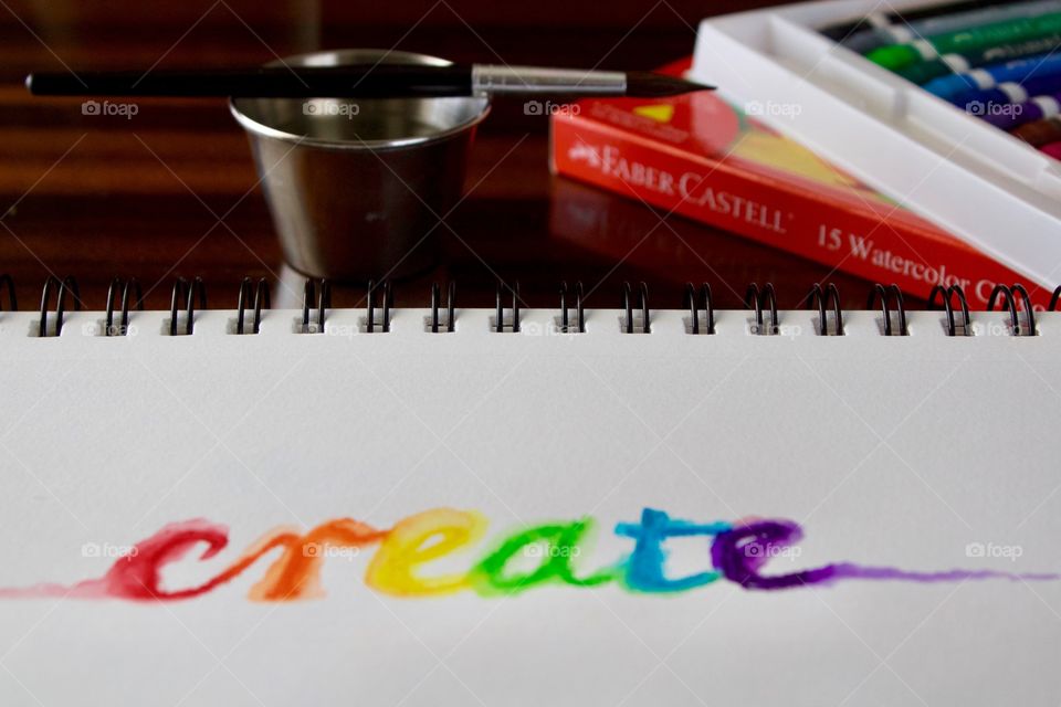 Colours of the World - watercolor crayons in white tray, brush and small stainless steel water container, product box, and the word “create” on mixed media wire-bound paper