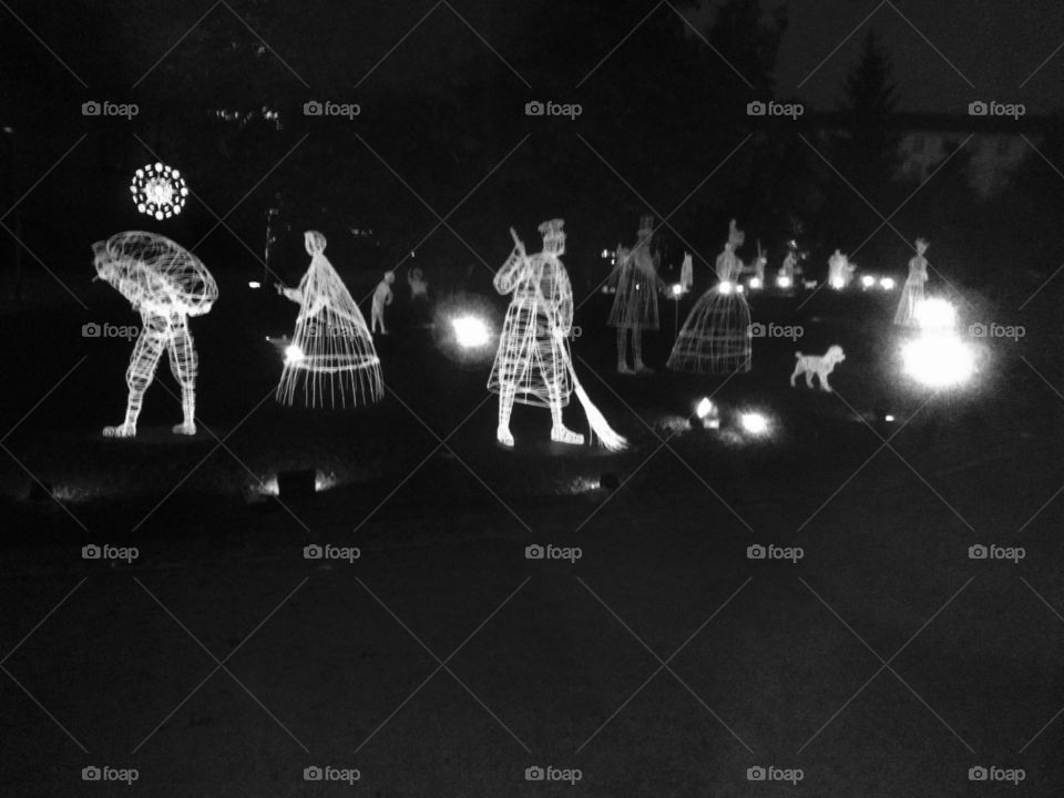 In a dark night a park is full of artworks, luminous iron wire figures, a man pack on his back, person has a broom in the hands, dog running around and spotlight further