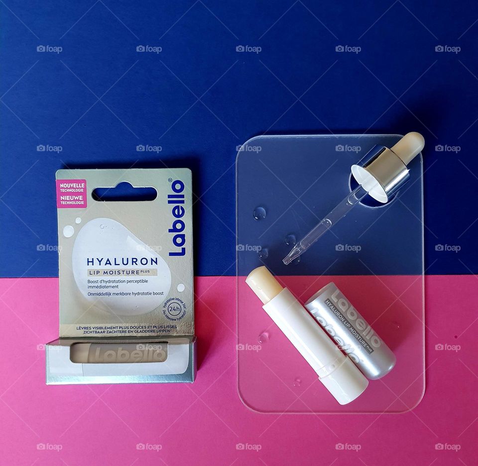 Flatlay on pink and navy blue papers of Labello Hyaluron lip moisture in its box and another one placed open on a small transparent plate with dropper and water drops