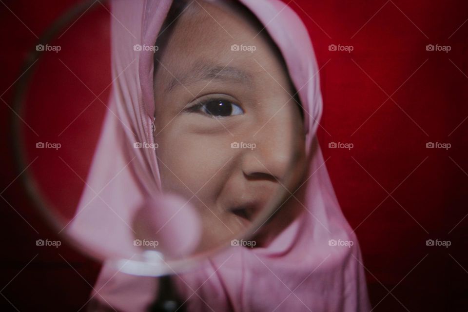 A little girl in a pink hijab, with a happy expression holding a magnifying glass to her face.