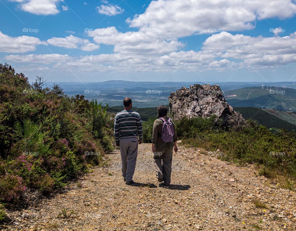 2 hikers trek along a stoney track, beyond them is a large rock and hills and valleys are in the far distance