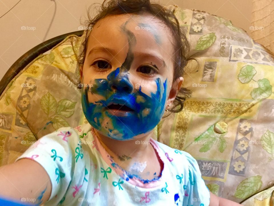 A little girl with messy paint color on face sitting on sofa