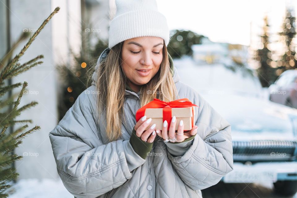Young happy woman blonde girl in warm clothes with gift box in hands near retro car decorated Christmas tree in winter street