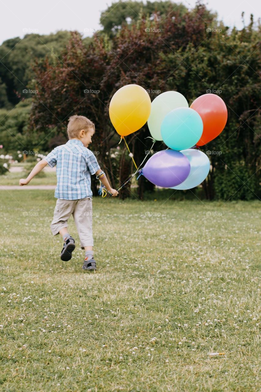 A little blond boy in a white T-shirt running on a green field, holding a bunch of colorful balloons in his hand, back view 