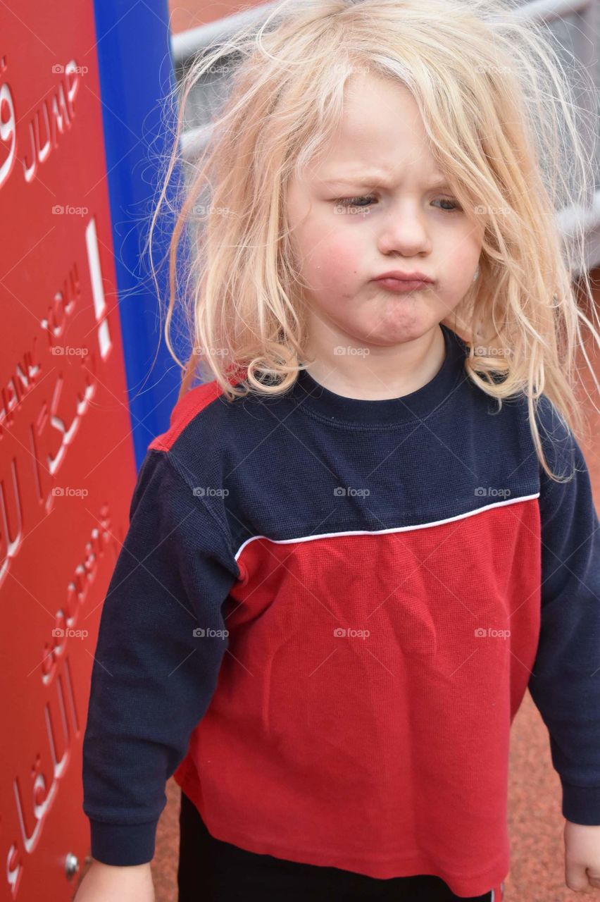 Pouting, toddler boy playing at the playground. There's red in the background. Little boy has long blonde hair.