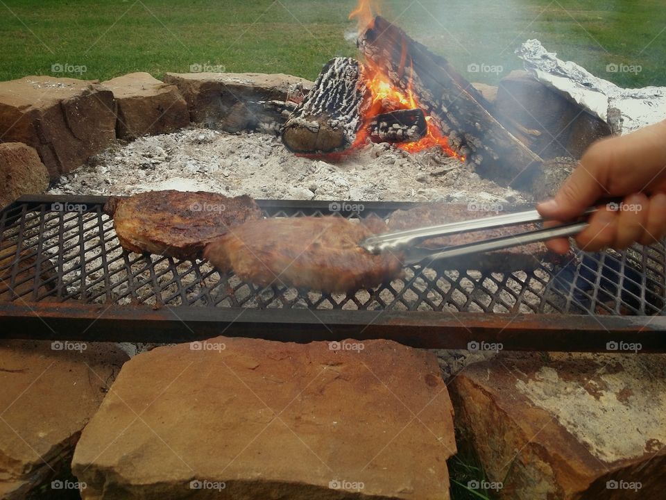 Cooking steaks over coals in an open fire pit with wood burning in the background and a man's hand flipping them with tongs