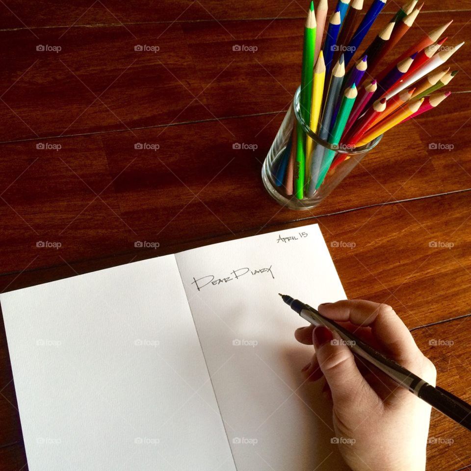 A woman's hand writing an entry in her journal with colorful pencils in frame on wooden surface
