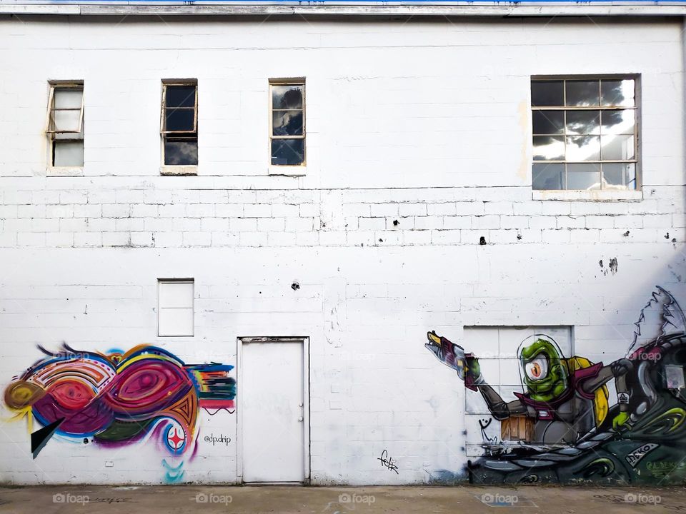 Street art on an abandoned old white rectangular cinder block style building with several old style rectangular windows and doors.