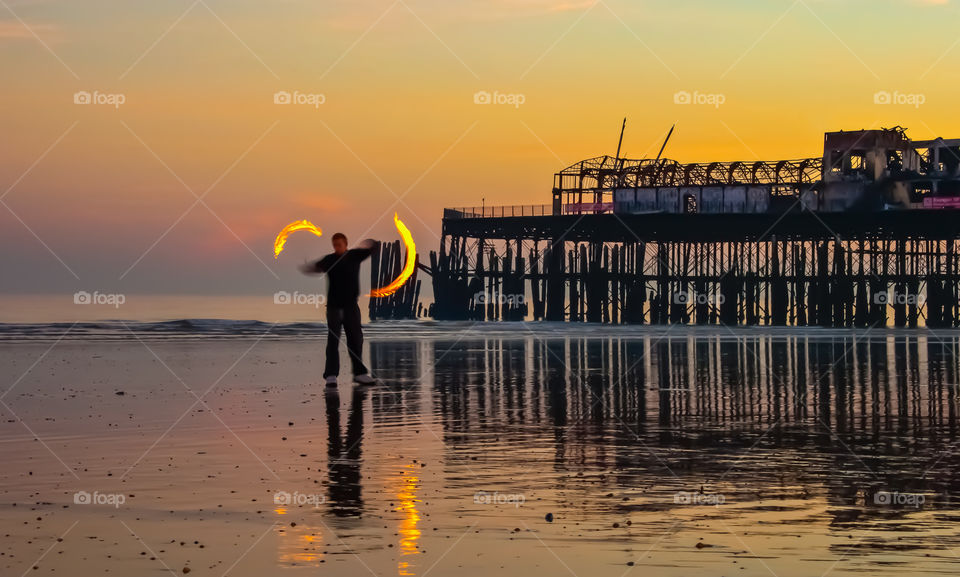 A man spins fire poi on the beach in front of the burnt remains of Hastings pier at sunset, UK 2010