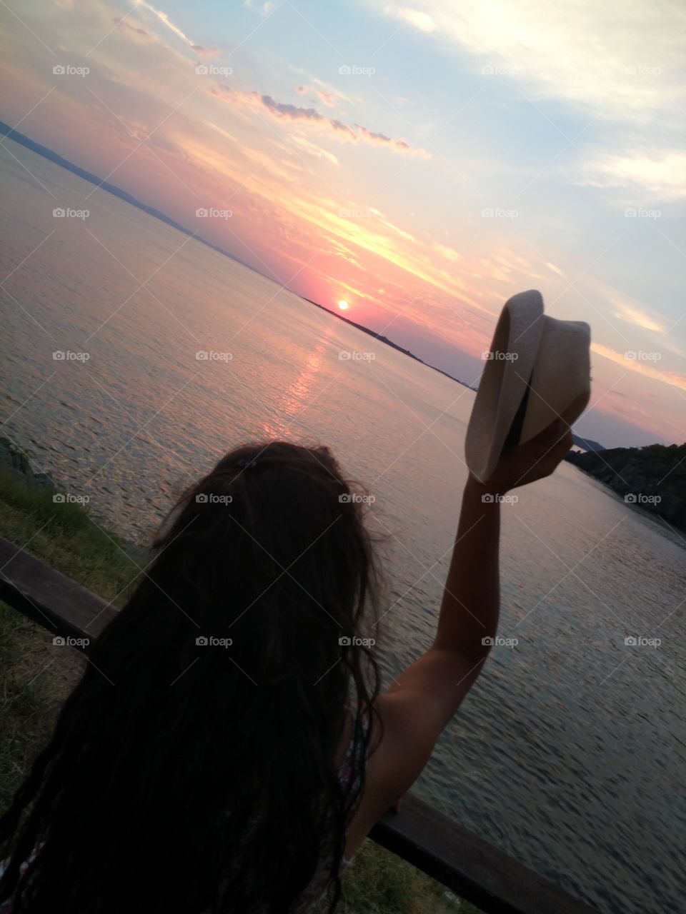 Brunette girl in the sunset holding her hat with her right hand, by the sea of summer in Northern Greece