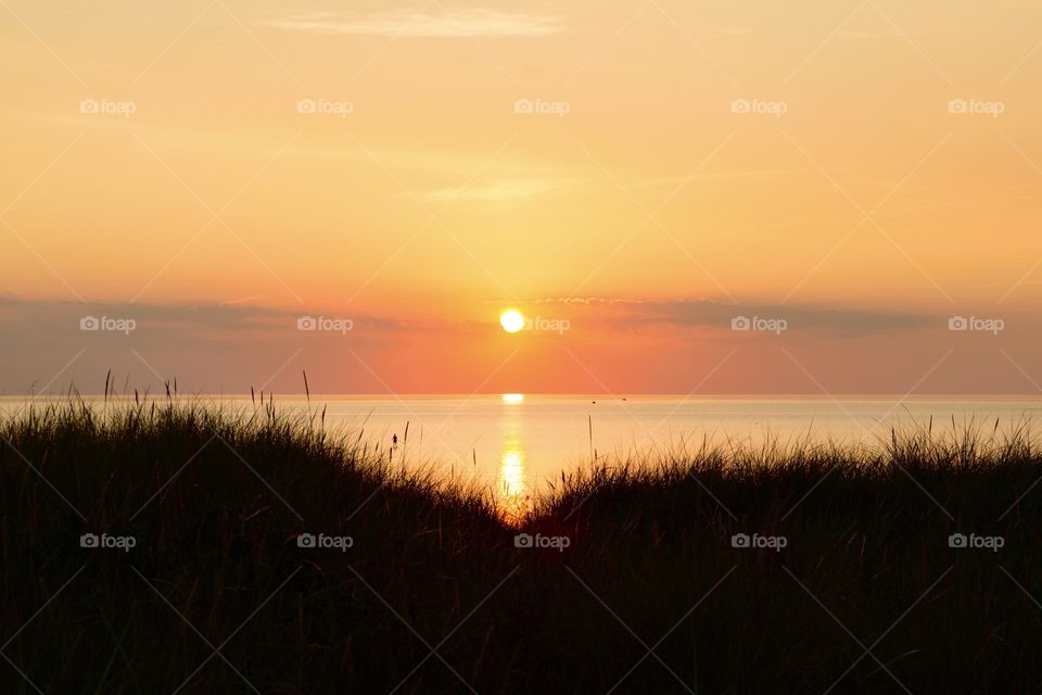 Beautiful orange colored sunset by the calm ocean, silhouette of sand dunes with grass in the foreground 