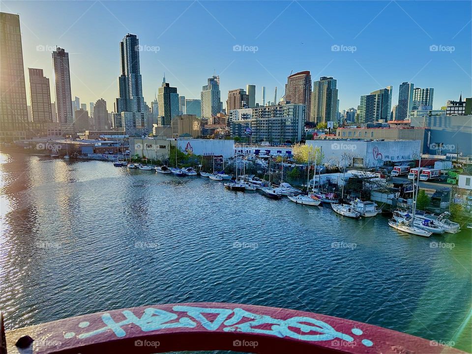 This is beautiful “Newtown Creek” seen from the “Pulaski Bridge” that connects “Greenpoint”, Bklyn to LIC, Queens with all its boats. The graffiti covered red metal railing encloses one of the main viewing platforms. 2024. Hypnotic Productions