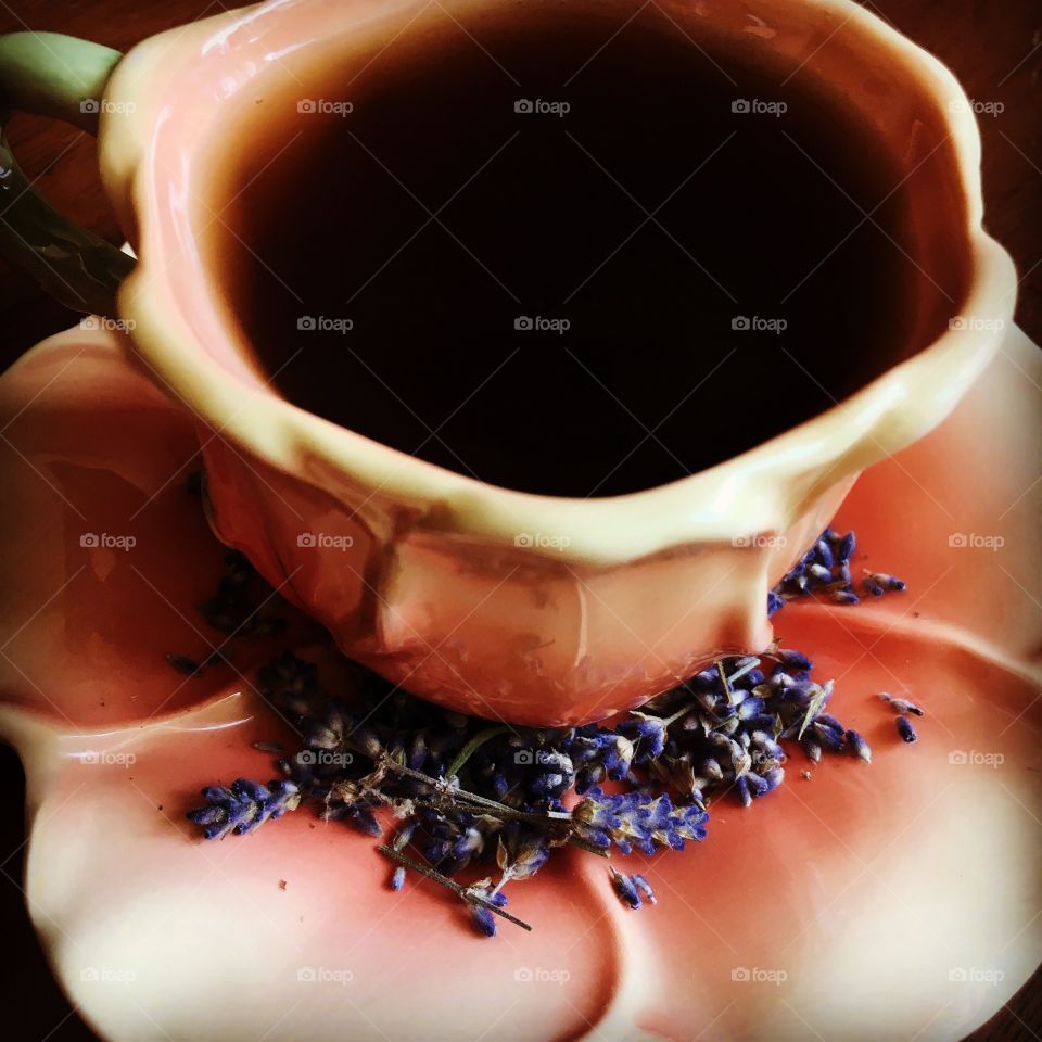 A dark cup of tea in a flower petal tea cup surrounded by bright and fragrant dried lavender flowers. 