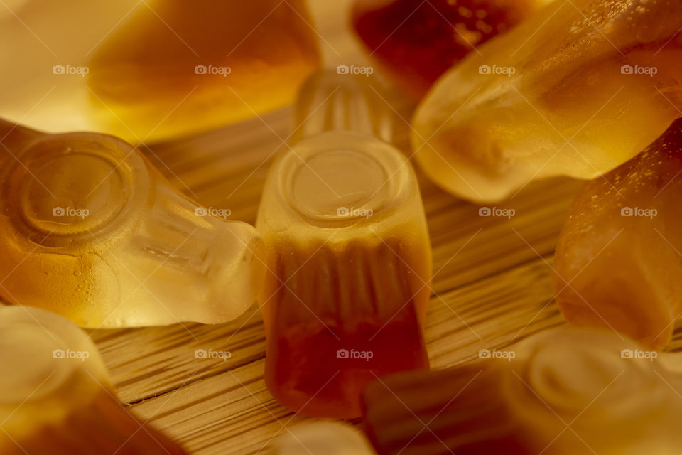 A macro portrait of a piece of candy shaped like a bottle of cola, surrounded by similar pieces of candy on a wooden table.