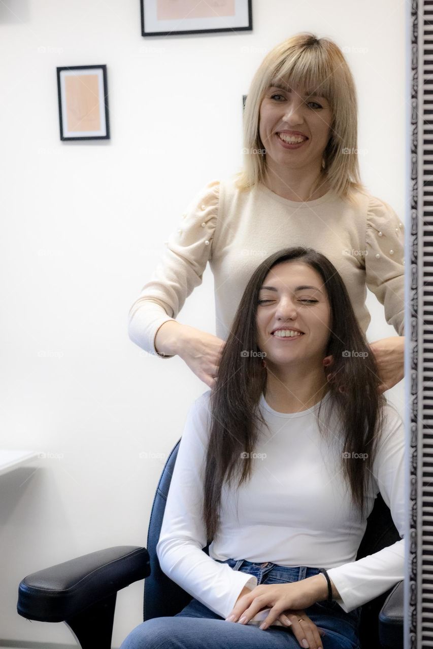 Portrait of beautiful girls blonde hairdresser showing the beauty and health of hair after styling and happy wide-smiling brunette clique sitting in a chair in the hairdressing salon,reflecting in the mirror,side view. The concept of happy people, ha