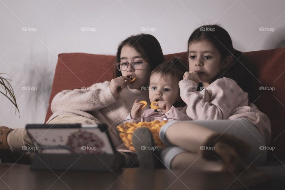 Portrait of three beautiful caucasian girls sisters are sitting on a sofa with a transparent glass dish with corn rings and enthusiastically watching a cartoon on a tablet standing on a coffee table in the room in the evening, close-up side view. Fam