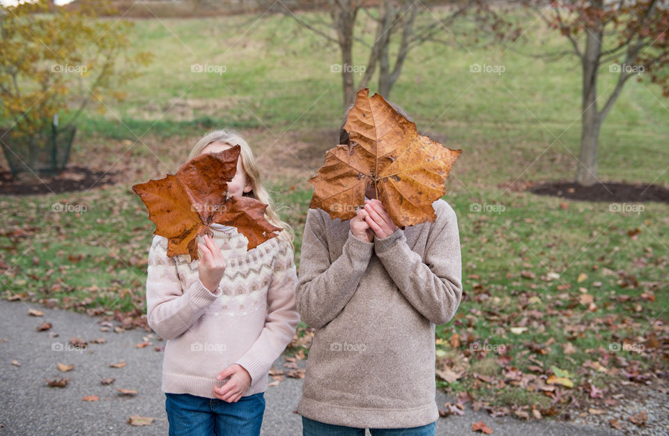 Two young sibling children holding large maple leaves over their faces outdoors during the fall