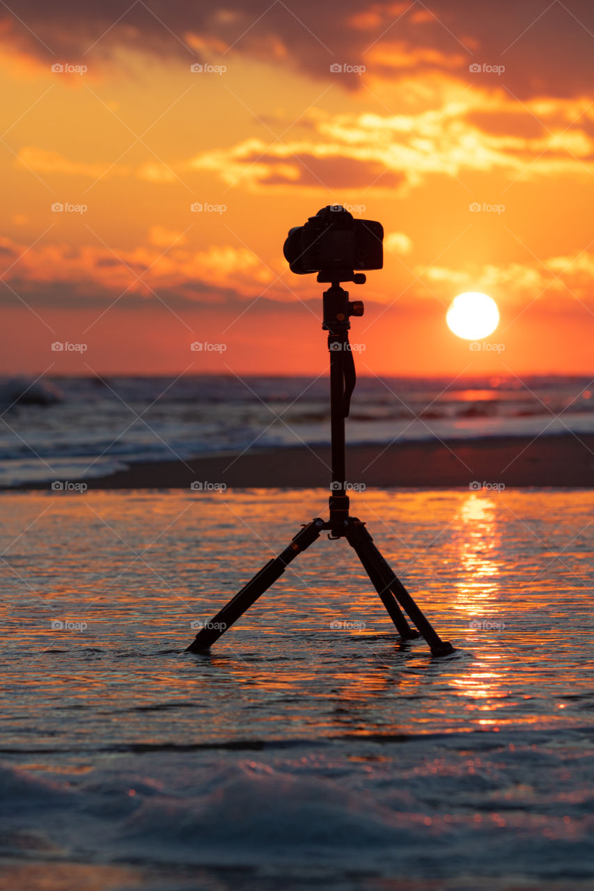 Selective focus on a DSLR camera capturing a beautiful sunset on a beach, as waves crash around the tripod and the surf is in the background.