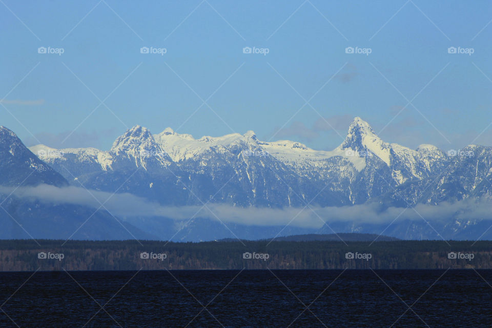 Another rare but welcome sunny winter day in Beautiful British Columbia. This shot is the Mainland mountains viewed from Vancouver Island. There is a wisp of  clouds over the foothills and the dark blue ocean of the Salish Sea in the foreground. 