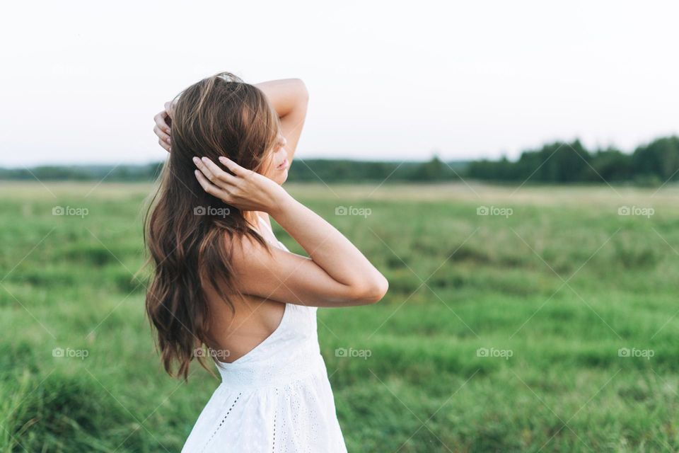 Young beautiful carefree long hair woman in white dress in sunset field, view from back. Sensitivity to nature concept